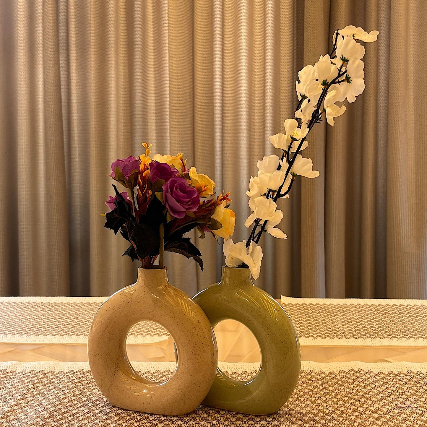 Two decorative vases with flowers on a tablecloth against a curtain background