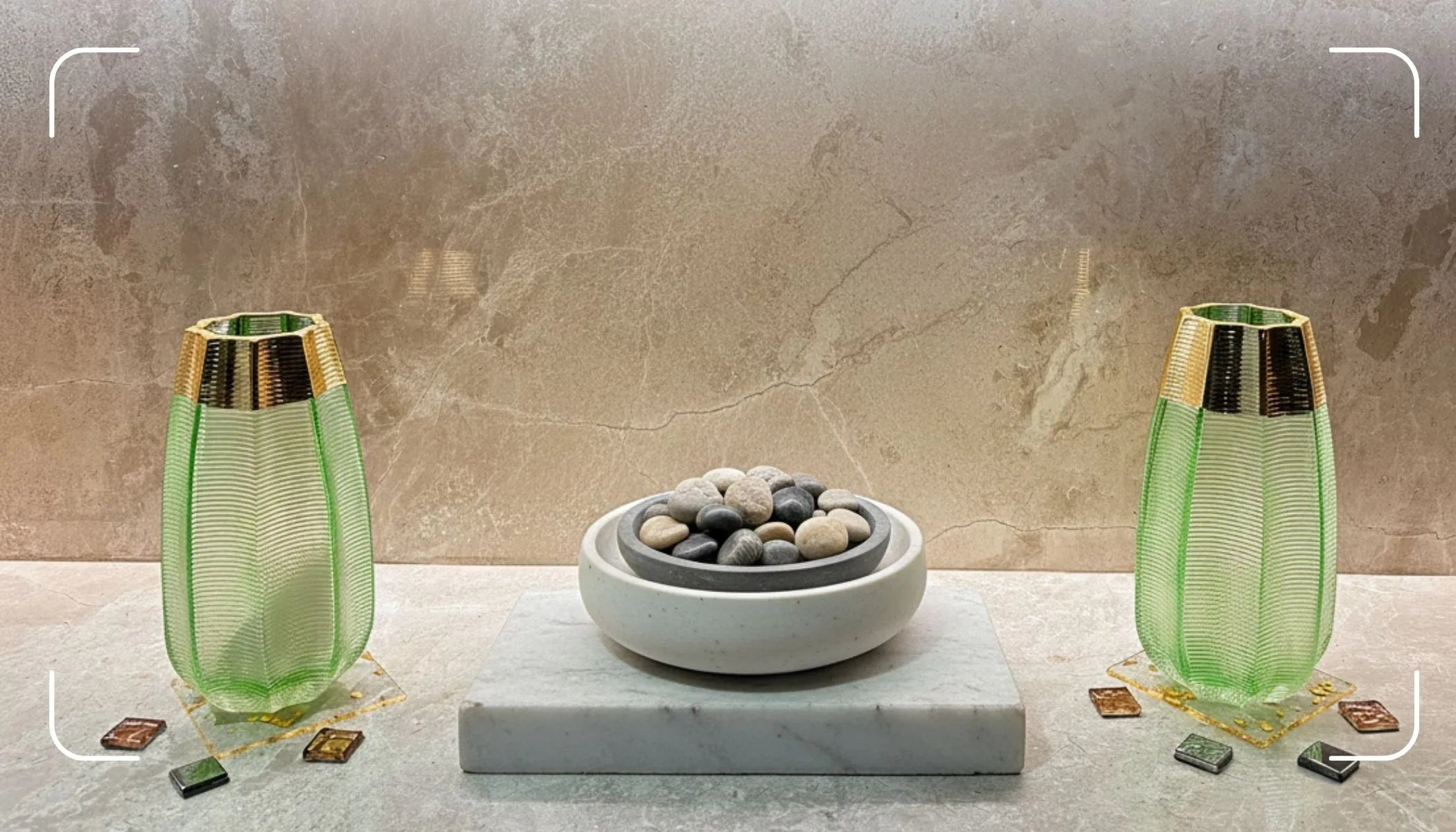 Two green glass vases with gold bases on a marble surface against a stone wall.