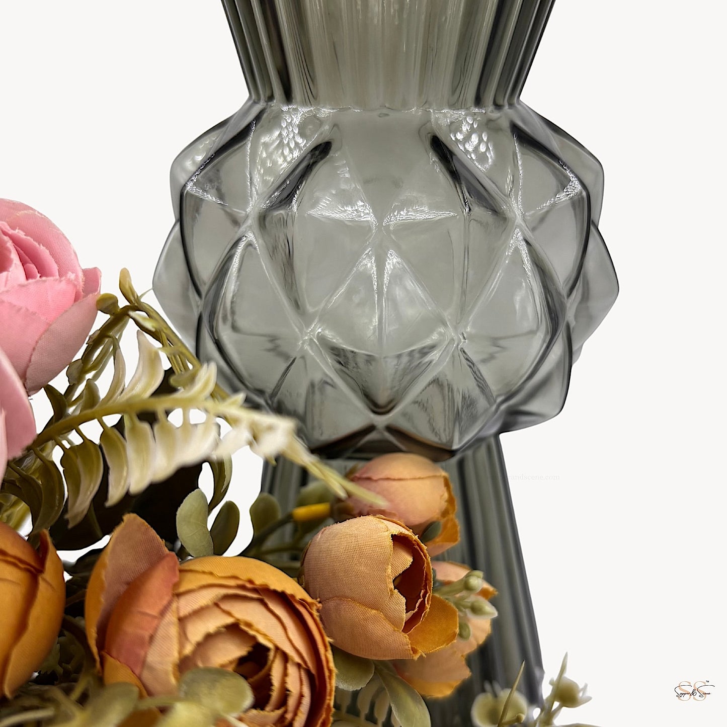 Close-up of a textured smoke-grey glass vase featuring a geometric diamond centerpiece and ribbed upper section, styled with soft pink and orange artificial flowers by Supper and Scene.