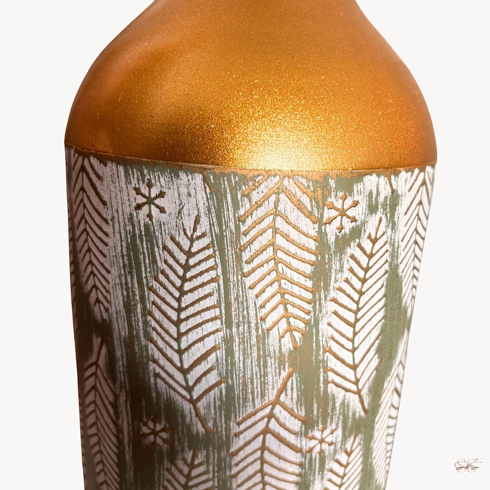 Close-up of a tall gold-and-green decorative metal vase with white leaf patterns and textured detailing by Supper & Scene.