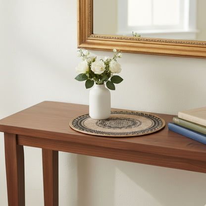 Round black woven placemat on a wooden surface with a vase and books in the background.