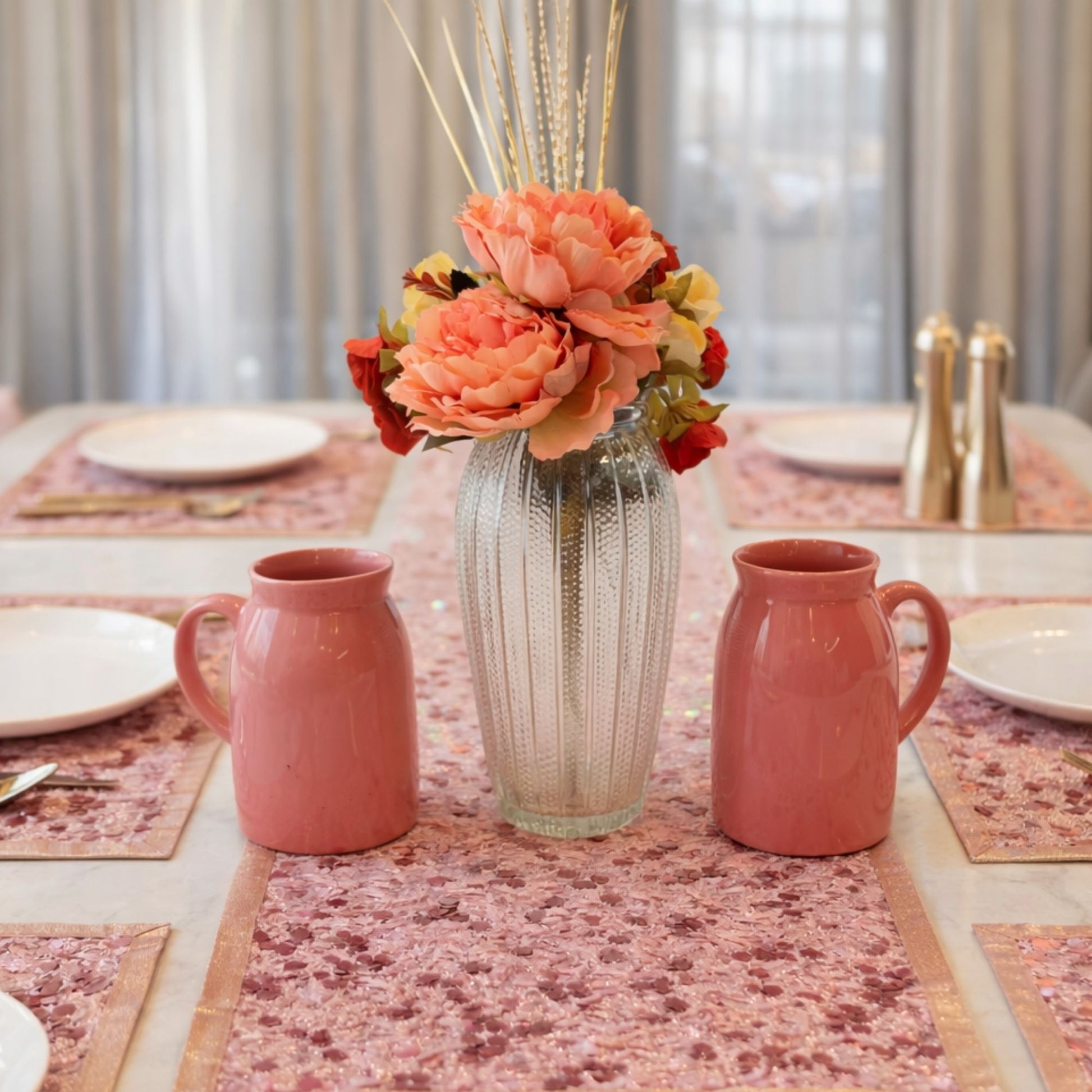 Decorative table setting with pink mugs, a vase with flowers, and patterned tablecloth.